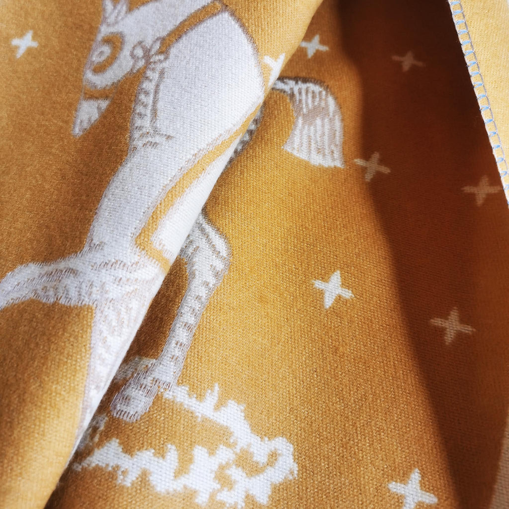 Detailed shot of the soft yellow knit fabric from a cashmere-feel winter scarf showing a white horse print and stitched edge. The fabric detail shows the fashion scarf is very soft and warm, and made with high-quality workmanship.