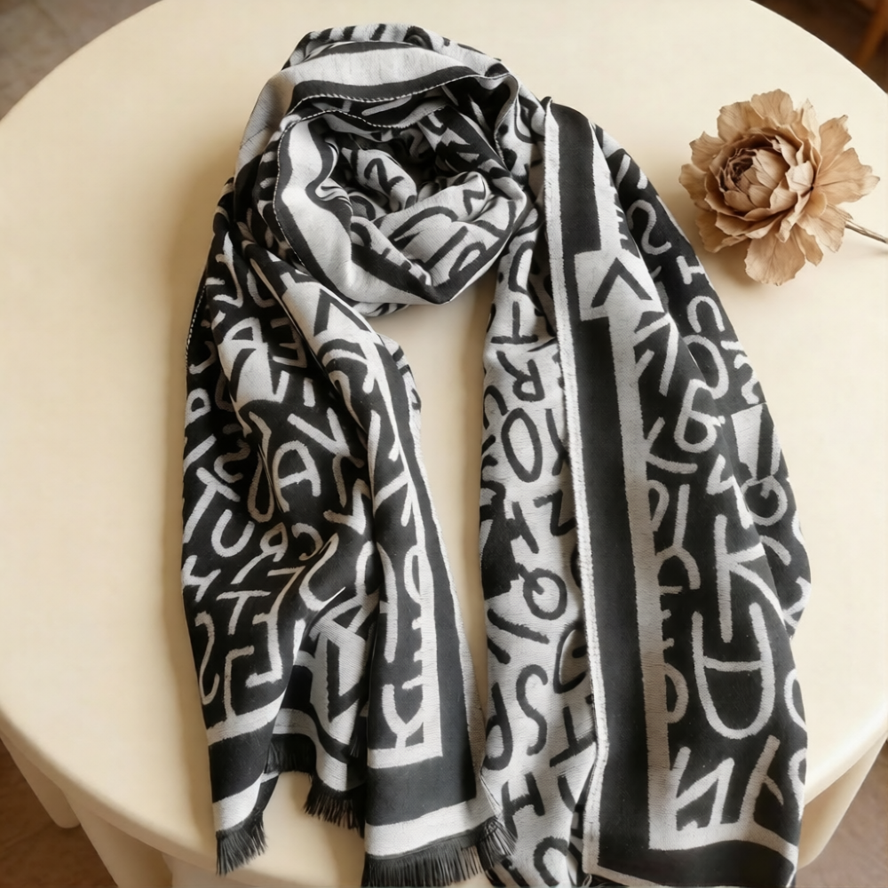 Women scarf with black and white letters pattern, styled on white table with a flower. The fashion winter scarf is large size, crafted from high-quality cashmere-feel fabric, looks very soft and warm.