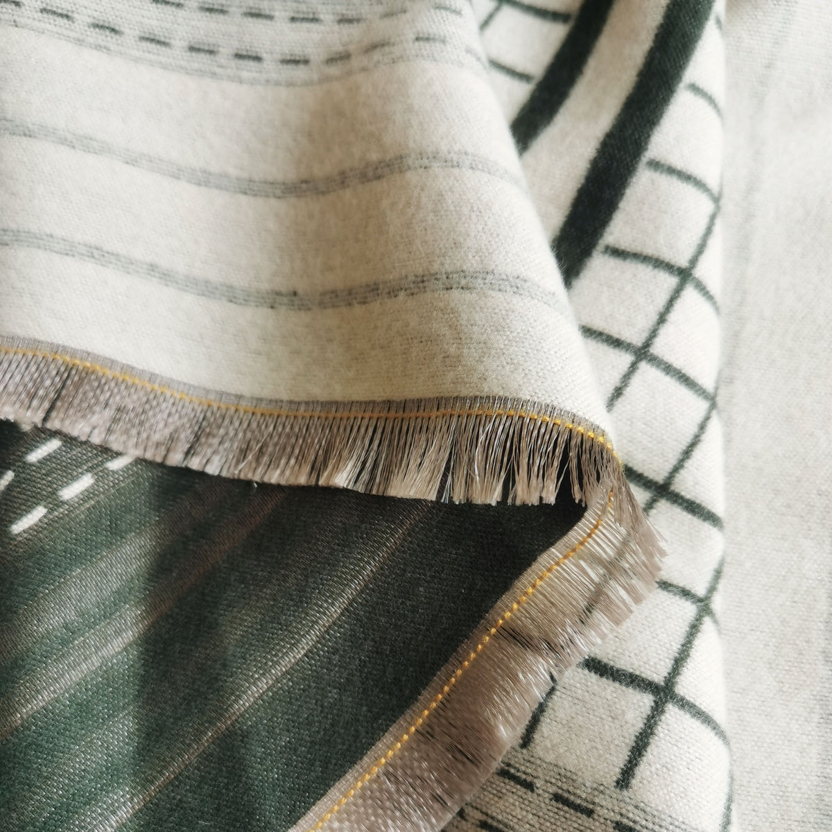 Close-up of a winter scarf fabric showing woven texture on dark green front and cream reverse side with fringe and yellow stitch edge. Exquisite details are showing this unisex scarf is crafted from cashmere-feel fabric with high-quality workmanship