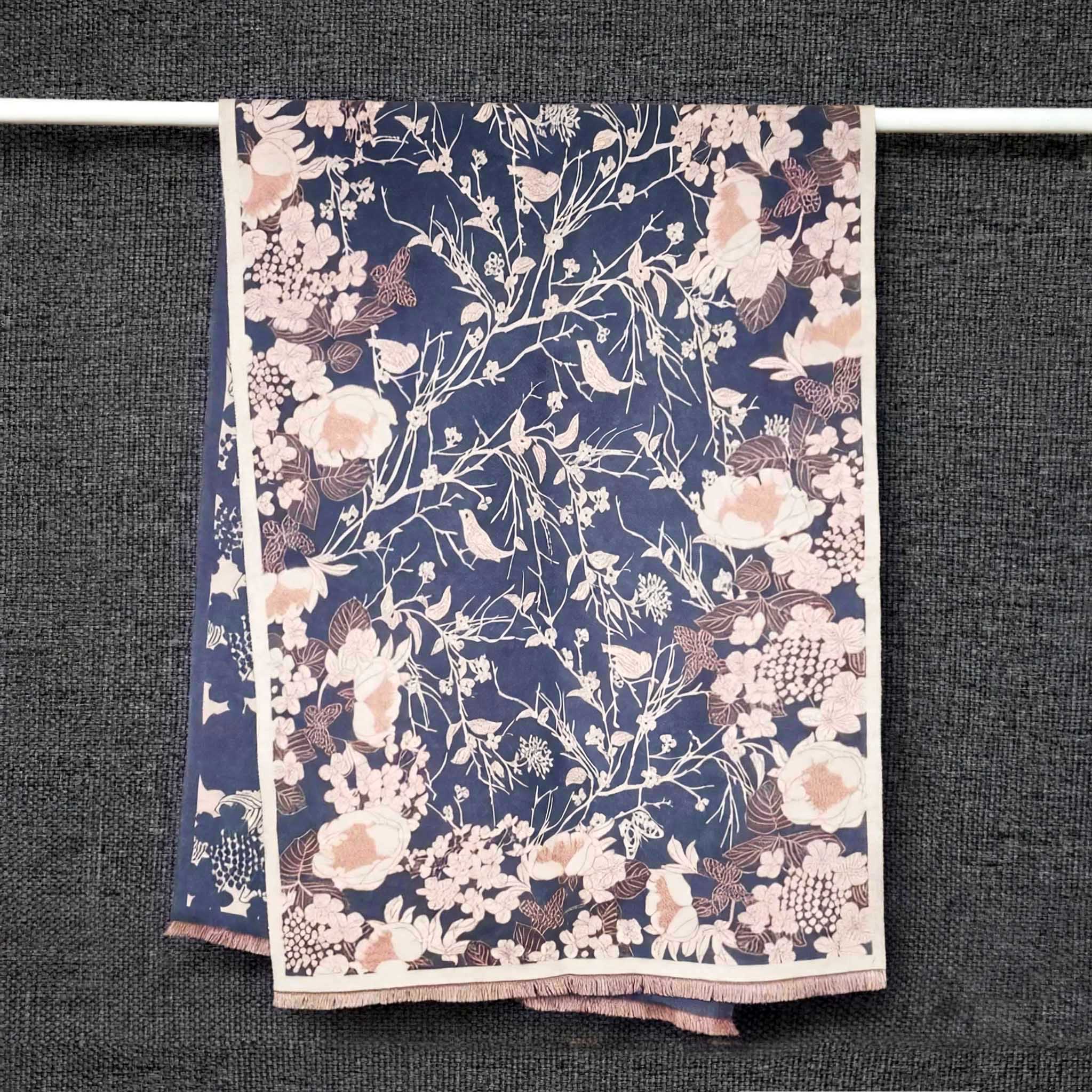 Full view of cashmere-feel winter women scarf in dusty blue hanging on display rail showing birds on branches and butterflies pattern with cream border and pink fringe